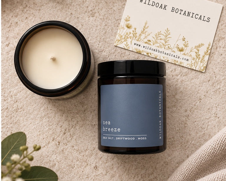 A dark glass candle jar labeled 'sea breeze' by Wildoak Botanicals, shown next to an open candle jar with cream wax and a branded card, styled on a textured beige surface with green leaves and a linen cloth.