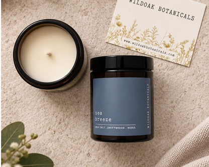 A dark glass candle jar labeled 'sea breeze' by Wildoak Botanicals, shown next to an open candle jar with cream wax and a branded card, styled on a textured beige surface with green leaves and a linen cloth.
