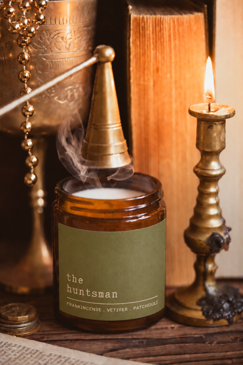 An amber glass jar candle labeled 'the huntsman' with a green label, shown lit with smoke rising from the top. The candle is styled on a wooden surface with a brass incense burner, a burning candlestick, books, and beads in a warm, moody setting.