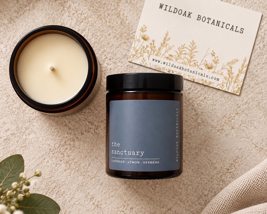 A dark amber glass candle jar with a slate blue label reading 'the sanctuary' is styled on a textured beige surface next to an open candle jar, a card for Wild Oak Botanicals, and green leaves.