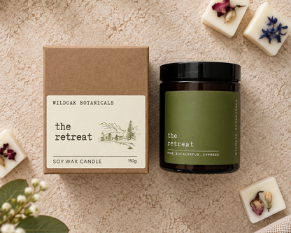 A soy wax candle by Wild Oak Botanicals titled 'the retreat', shown with its brown kraft paper box and an amber glass jar with a dark green label. The product is styled in a flat lay on a textured beige linen background, surrounded by dried flowers in wax melts and green leaves.