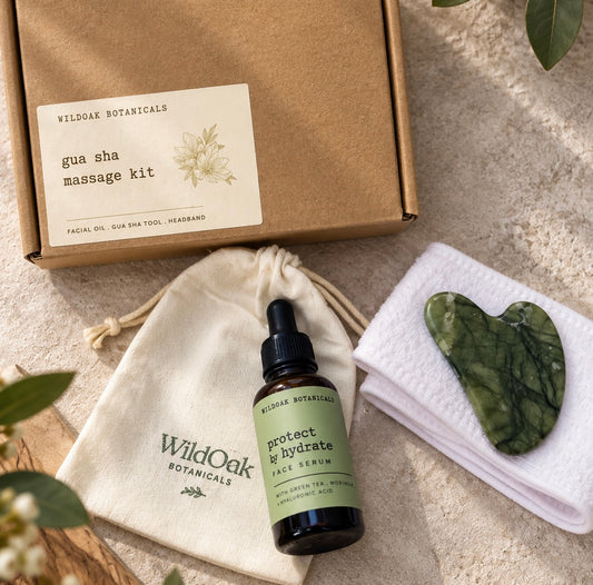 Wild Oak Botanicals gua sha massage kit arranged on a neutral surface, including a kraft gift box, cream drawstring pouch, small amber dropper bottle labeled face serum, folded white towel, and green marbled stone tool with a heart-shaped cutout.