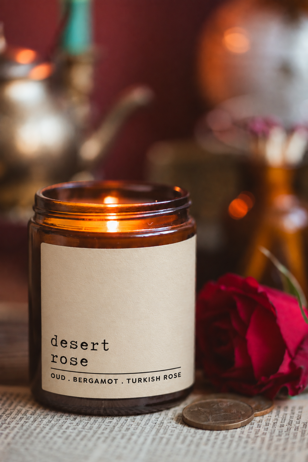 An amber glass jar candle labeled 'desert rose' with a lit flame, styled on a textured surface with a red rose and coin beside it, against a warm blurred background.