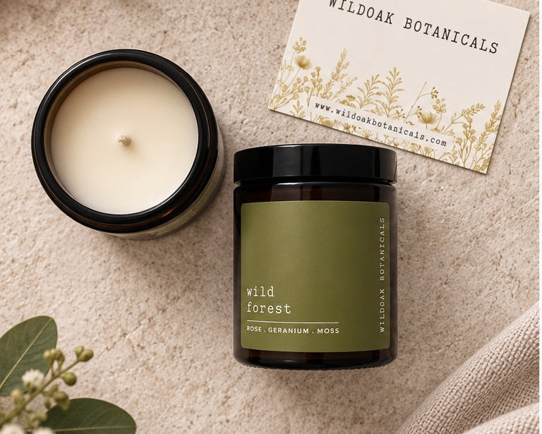 A jar candle and an open candle jar with cream wax are arranged on a textured beige surface with greenery and a card reading Wildoak Botanicals. The jar has an olive green label and a black lid.