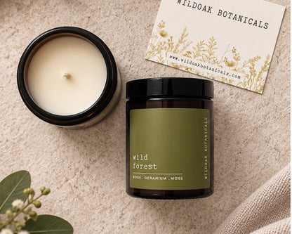 A jar candle and an open candle jar with cream wax are arranged on a textured beige surface with greenery and a card reading Wildoak Botanicals. The jar has an olive green label and a black lid.