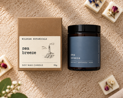 Wild Oak Botanicals 'sea breeze' soy wax candle in an amber glass jar with a black lid, displayed next to its brown kraft paper box on a beige textured blanket with decorative botanicals and wax melts.